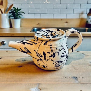 May include: Cream-colored ceramic teapot with a black splatter design. The teapot has a curved handle, a spout, and a lid. The teapot is sitting on a wooden surface.