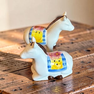 May include: Two white ceramic donkey salt and pepper shakers. Each donkey features a brown mane and a painted blanket on its back with a yellow center, pink floral details, and blue striped borders. The shakers have small holes on the blankets.