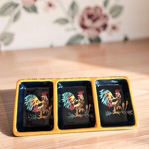 May include: A rectangular ceramic serving tray with three separate compartments. Each section features a glossy dark blue interior with a hand-painted rooster illustration in shades of red, yellow, and blue. The outer rim is a solid mustard yellow.
