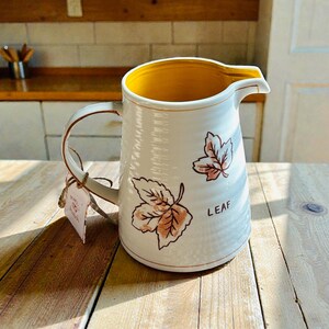 May include: White ceramic pitcher with a yellow interior and a curved handle. The pitcher features two leaf illustrations in shades of brown and pink, with the word "LEAF" printed on the side. A tag is attached to the handle.