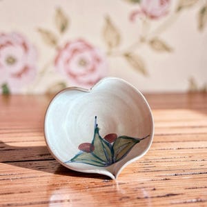 May include: A heart-shaped ceramic bowl with an off-white glaze. The interior features a hand-painted design of green leaves and small reddish-brown berries centered at the bottom. The bowl has a smooth, curved rim and a tapered base.