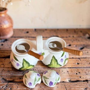 May include: A double ceramic condiment server with a central handle, featuring hand-painted green leaves and purple berry clusters. Two small metal spoons with wooden handles are inside, and two matching ceramic lids are placed in front.