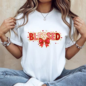 May include: White t-shirt with the word "BLESSED" in a decorative font. The letters are red, pink, and white, with heart and bow accents. The shirt is a casual, everyday wear item.