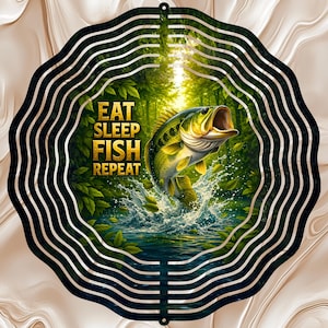 May include: A round metal wall art piece with a fishing theme. The artwork features a leaping bass fish in a forest scene with the words "EAT SLEEP FISH REPEAT". The design is framed by a series of concentric wavy lines.