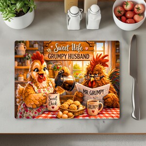 Peut inclure: Une planche &agrave; d&eacute;couper rectangulaire illustr&eacute;e de deux poulets dans une cuisine. Un poulet porte un tablier &agrave; fleurs et verse du caf&eacute; dans une tasse marqu&eacute;e Sweet Chick. L'autre porte des lunettes, lit un journal intitul&eacute; Mr. Grumpy et est assis pr&egrave;s d'une tasse marqu&eacute;e Grumpy. Le texte indique Sweet Wife Grumpy Husband.