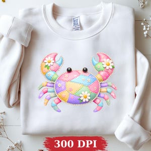 May include: A white crewneck sweatshirt featuring a central graphic of a crab with a patchwork design in pastel shades of pink, blue, yellow, and purple. The crab is decorated with small white flowers and pearl-like accents. Text reads 300 DPI.