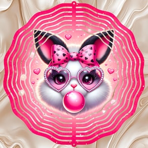 May include: A circular metal wind spinner featuring a vibrant pink scalloped border and a central illustration of a white bunny with black-tipped ears. The bunny wears a pink bow with black hearts and heart-shaped glasses, blowing a pink bubble.