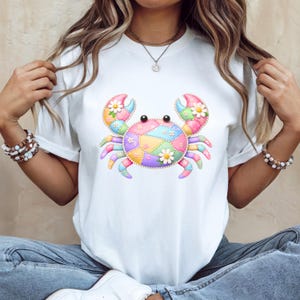 May include: White t-shirt featuring a colorful crab design. The crab is made of patchwork in pastel colors, with daisy accents on the claws. The design is centered on the shirt, creating a playful and summery aesthetic.