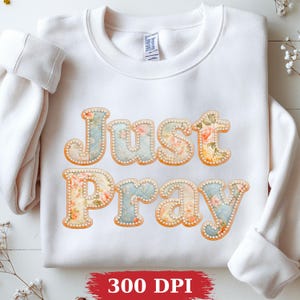 Peut inclure: Un sweat-shirt blanc à manches longues avec le texte "Just Pray" en grandes lettres style bloc. Les lettres présentent un motif floral vintage dans des tons bleu, crème et pêche, souligné par une bordure décorative de petites perles blanches.