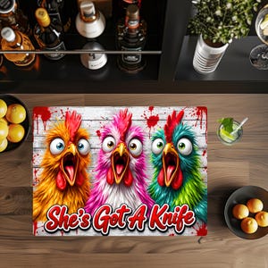 Puede incluir: Una alfombrilla rectangular con tres pollos de dibujos animados con ojos muy abiertos y picos abiertos en colores naranja, rosa y verde azulado sobre un fondo de tablones de madera blanca con salpicaduras rojas. Texto She's Got A Knife en rojo.