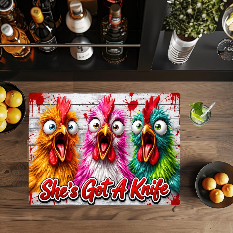 Puede incluir: Una alfombrilla rectangular con tres pollos de dibujos animados con ojos muy abiertos y picos abiertos en colores naranja, rosa y verde azulado sobre un fondo de tablones de madera blanca con salpicaduras rojas. Texto She's Got A Knife en rojo.