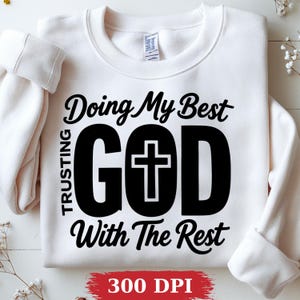 May include: White sweatshirt with the text "Doing My Best, Trusting God With The Rest" in black. The word "GOD" is large and centered, with a cross inside the letter "O". The bottom of the sweatshirt has the text "300 DPI" in red.