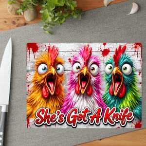 Puede incluir: Una tabla de cortar rectangular con un gr&aacute;fico de tres pollos de ojos muy abiertos y gritando en amarillo, rosa y verde azulado sobre un fondo de tablones de madera blanca con salpicaduras de pintura roja. Texto "She's Got A Knife" en rojo.