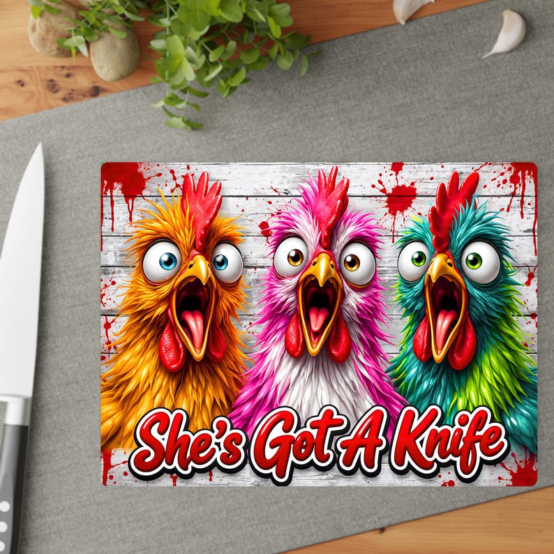 Puede incluir: Una tabla de cortar rectangular con un gr&aacute;fico de tres pollos de ojos muy abiertos y gritando en amarillo, rosa y verde azulado sobre un fondo de tablones de madera blanca con salpicaduras de pintura roja. Texto "She's Got A Knife" en rojo.