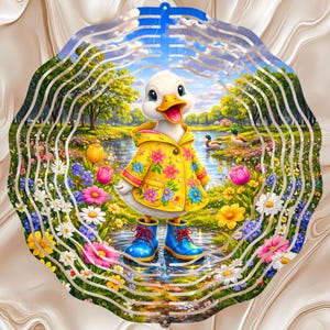 May include: A circular metal wind spinner featuring a colorful illustration of a white duckling wearing a yellow floral raincoat and blue rain boots, standing in a puddle surrounded by vibrant wildflowers, green trees, and a pond with two ducks.
