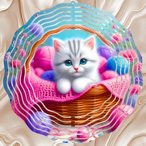 May include: A fluffy white kitten with gray stripes and bright blue eyes sits in a woven basket filled with pink, purple, and blue yarn balls. The basket rests on a pink knitted blanket, surrounded by decorative flowers.