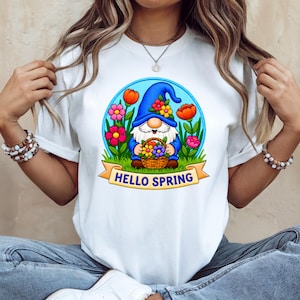 May include: White t-shirt featuring a colorful graphic of a gnome holding a basket of flowers, surrounded by flowers and the text "HELLO SPRING". The gnome wears a blue hat and is set against a blue background.