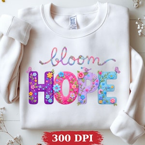 May include: A cream-coloured crewneck sweatshirt featuring the word bloom in a multicoloured script font above the word HOPE in large, block letters. The letters are filled with colourful floral patterns, small birds, and butterflies.