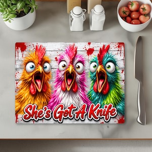 Puede incluir: Una tabla de cortar rectangular de cristal con tres pollos de dibujos animados en amarillo, rosa y turquesa con ojos grandes y picos abiertos. Fondo de madera blanca con salpicaduras rojas y el texto She's Got A Knife.