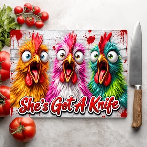 Puede incluir: Una tabla de cortar rectangular con un gr&aacute;fico de tres pollos de ojos muy abiertos y gritando en colores amarillo, rosa y verde azulado sobre un fondo de madera blanca con salpicaduras rojas y el texto She's Got A Knife en letras rojas.