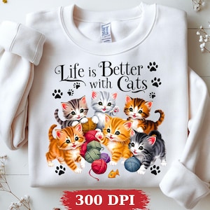 May include: A white crewneck sweatshirt featuring a graphic of six kittens playing with colorful yarn balls. The text "Life is Better with Cats" is printed in black above the kittens, surrounded by small black paw prints. A small orange fish is visible.