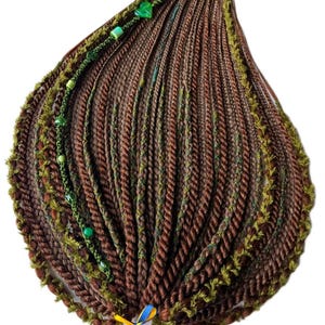 May include: A set of brown synthetic hair extensions styled in multiple twists and braids, accented with green yarn, small green beads, and a heart-shaped charm. The arrangement is secured at the base with a small blue and yellow ribbon tie.