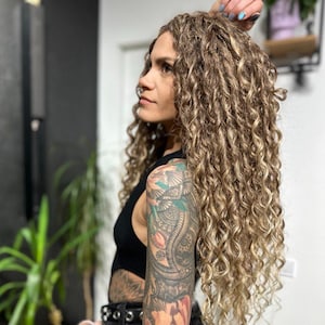 Op de afbeelding: Een vrouw met lang, krullend, lichtbruin haar. Ze draagt een zwarte top en een zwarte broek. Haar arm is bedekt met kleurrijke tatoeages, waaronder bloemen- en paisley-ontwerpen. Ze draagt een zwart horloge en een zwarte riem.