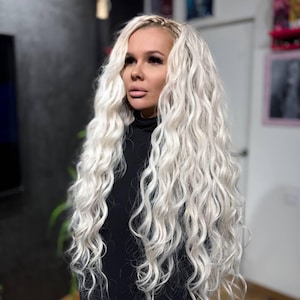 Op de afbeelding: Een vrouw met lang, golvend, platina blond haar. Ze draagt een zwarte coltrui. Het haar is gestyled met een zijvlecht en valt in volumineuze krullen rond haar schouders.