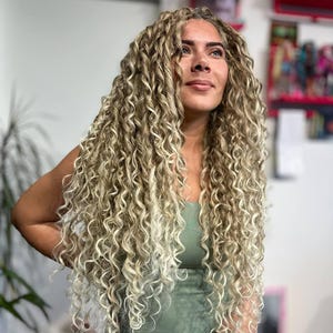 Puede incluir: Extensiones de cabello rubio rizado y largo caen en cascada. El cabello tiene un aspecto natural y texturizado con diferentes tonos de rubio. La persona lleva un top verde y una parte inferior blanca. El fondo es una pared de color claro con algo de decoración.