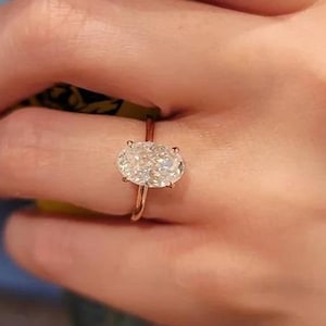 Può includere: Anello in oro rosa con un grande diamante taglio ovale. Il diamante è il punto focale, incastonato su una fascia sottile. L'anello è mostrato su una mano, evidenziando la gemma scintillante.