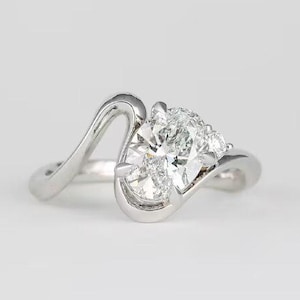 May include: A silver-toned ring featuring a central oval-cut clear gemstone held in a bypass-style setting. The band curves around the stone, which is accented by a smaller round-cut clear gemstone on one side, creating a fluid, asymmetrical design.