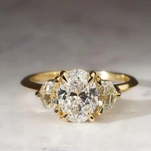 May include: A gold engagement ring with a large oval-cut diamond center stone, flanked by two smaller, rectangular diamonds. The ring's band is a simple, polished gold. The diamonds sparkle against a neutral background.