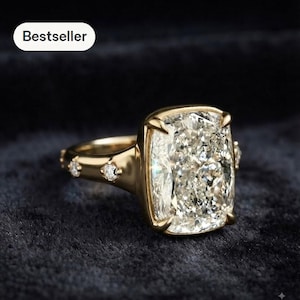 Op de afbeelding: Een gouden ring met een grote, rechthoekige cushion-cut steen die door vier pootjes wordt vastgehouden. De band is gemaakt van gepolijst goud en is versierd met kleine, ronde, heldere steentjes die in de zijkanten zijn ingelegd.