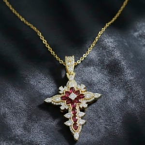 May include: A gold-colored pendant necklace featuring a cross design. The cross is adorned with red gemstones and clear crystals, set in a detailed, ornate pattern. The necklace hangs on a delicate gold chain against a dark background.