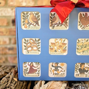 May include: A blue box with a red ribbon, featuring illustrated tiles with numbers and text. The tiles depict images like a partridge, turtle doves, gold rings, geese, swans, ladies dancing, lords, and pipers.