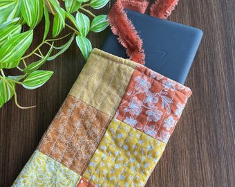 May include: A patchwork Kindle sleeve in shades of yellow, orange, and brown, with floral and polka dot patterns. A dark blue Kindle is partially visible inside. A rust-colored ribbon is tied at the top. The sleeve is on a wooden surface.