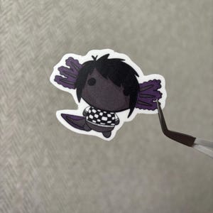 May include: A sticker of a cartoon axolotl character with black hair and a checkered shirt. The axolotl has purple gills and a tail. The sticker is held by tweezers against a grey background.