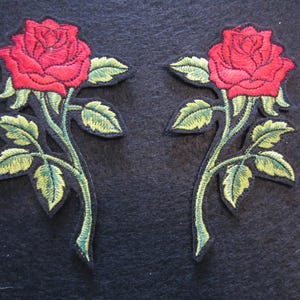 May include: Two embroidered rose patches. Each patch features a vibrant red rose with detailed petals, green leaves, and stems. The patches are set against a dark background, highlighting the colorful floral design. Ideal for fabric embellishment.