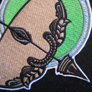 May include: Embroidered patch with a black and white design of an arrow and a bow, set against a green circle with a white border.