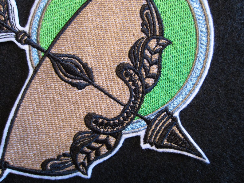 May include: Embroidered patch with a black and white design of an arrow and a bow, set against a green circle with a white border.