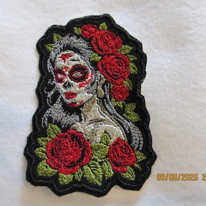 May include: An embroidered patch featuring a person with sugar skull makeup, long grey hair, and red lips. The design includes vibrant red roses and green leaves surrounding the figure, all outlined with a thick black border on a white background.