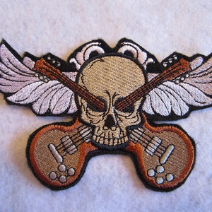May include: Embroidered patch featuring a skull with crossed guitars and wings. The skull is tan with black details, and the guitars are brown and tan. The wings are white with black outlines. The patch is designed for clothing or accessories.