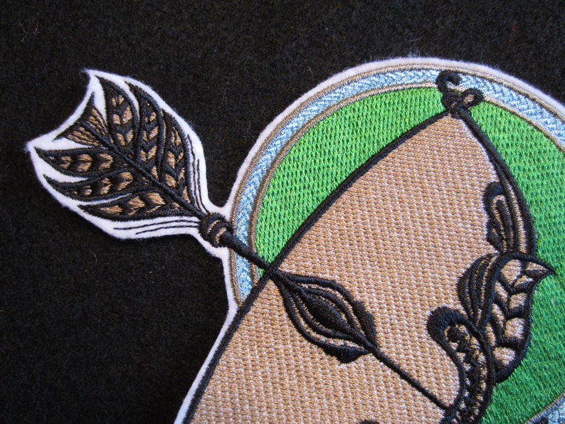 May include: Embroidered patch with a brown bow and arrow design. The arrow has a brown and black feathered fletching. The bow is surrounded by a green circle with a blue and white border.