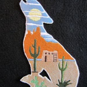 May include: Embroidered patch featuring a coyote silhouette with a desert landscape inside. The coyote is howling at the moon, which is depicted as a yellow circle. The desert landscape includes a cactus, a small building, and a ladder.