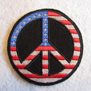 May include: A round embroidered patch with a peace sign design. The peace sign is outlined in white and filled with the colors of the American flag. The background is a white fabric.