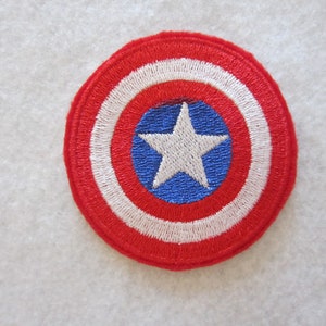 May include: Embroidered patch featuring a shield design. The patch has a red outer ring, followed by white and red concentric circles. A blue circle in the center contains a white five-pointed star. This patch is likely inspired by a superhero.