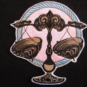 May include: Embroidered patch featuring the astrological symbol for Libra, a set of scales with ornate details. The scales are brown and the background is pink with a blue and white border.