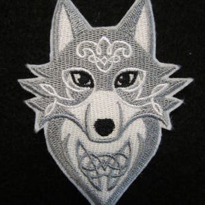 May include: A silver embroidered wolf head patch with a Celtic knot design. The wolf's eyes are black and the mouth is open in a snarl.