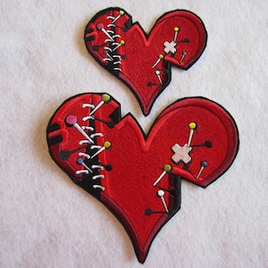 May include: Two red felt heart-shaped patches with black stitching and colorful pins. The hearts are broken and stitched back together.