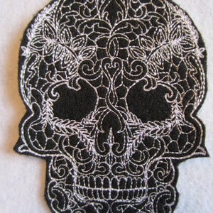 May include: Black felt skull with white embroidery in a detailed floral pattern. The skull has a traditional sugar skull design.